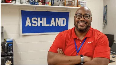 Athletic Director Kevin Johnson wearing a red polo shirt in front of a blue "Ashland" pennant
